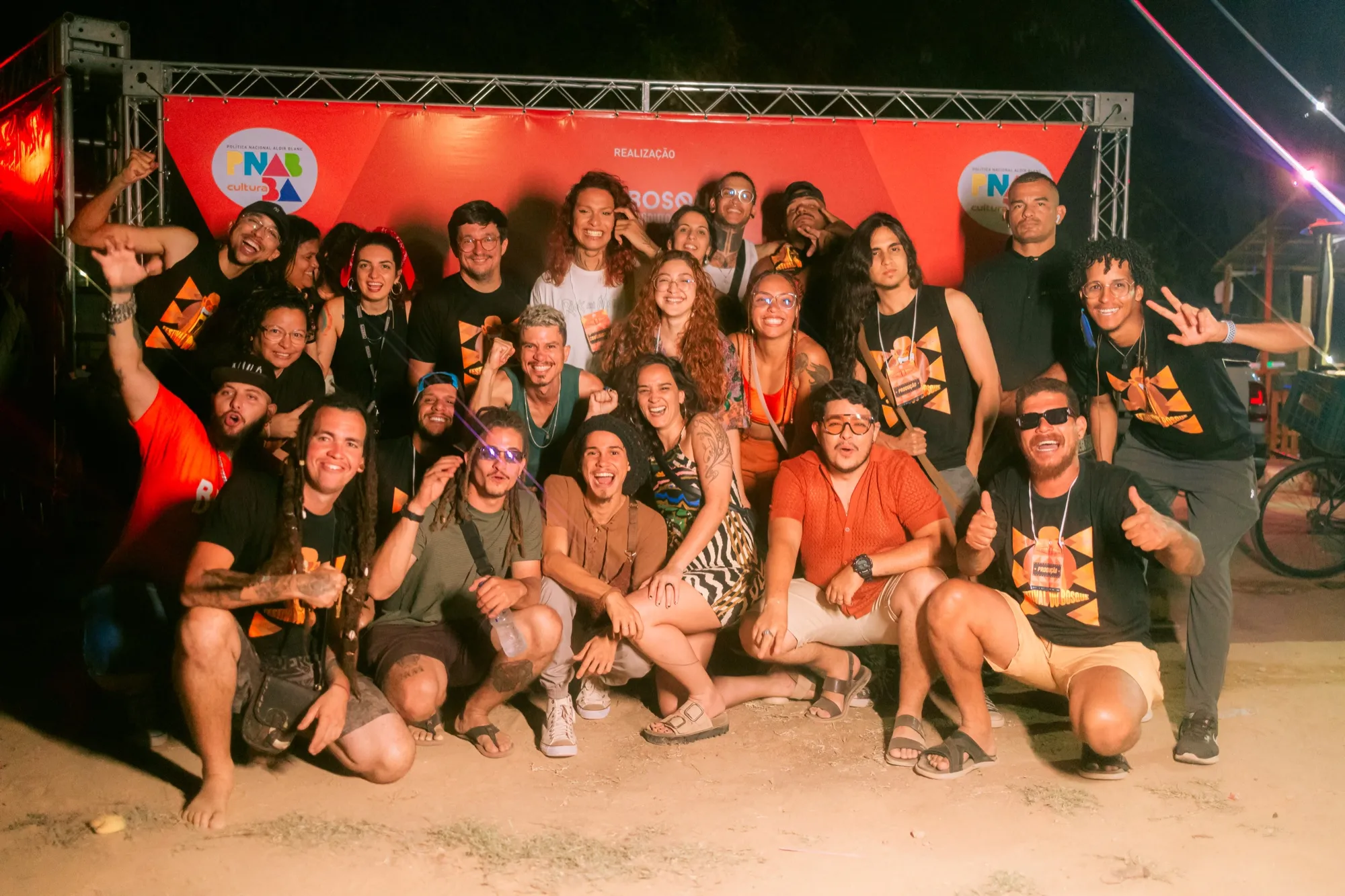 Equipe da Bosque Produtora reunida e sorrindo diante do painel de realização do Festival do Bosque, na terceira edição, com camisetas da produção